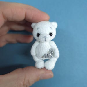 Peut inclure: Un petit ours en peluche blanc avec des yeux noirs et une décoration en paillettes argentées sur la poitrine. L'ours est fait d'une matière douce et duveteuse et a un design simple et attachant. L'ours est tenu dans une main sur fond bleu.