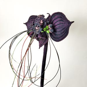 Puede incluir: Una flor de ganchillo púrpura oscuro hecha a mano con hojas grandes y texturizadas y detalles intrincados. La flor presenta flores más pequeñas y agrupadas en tonos de púrpura, gris y rosa, con delicados acentos de alambre que se extienden hacia afuera. El tallo es púrpura oscuro.