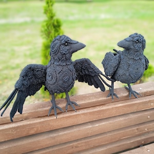 May include: Two black crochet raven sculptures with detailed wing and body patterns. One raven has its wings spread, while the other has its wings folded. The birds are perched on a wooden bench, set against a blurred green background.