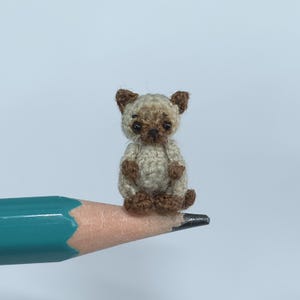 May include: A tiny crocheted Siamese cat, with a beige body and brown details on its ears, paws, and face. The miniature cat is sitting on a pencil tip, highlighting its small size and intricate design. The pencil is a standard size.