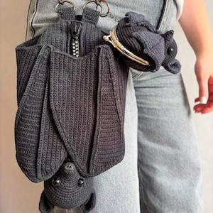 May include: A dark grey crochet bat-shaped bag with a zip pocket and a small coin purse. The bag has a chain strap and is designed to be worn on the hip. The bat's wings are spread, and it has small, round eyes.