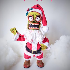 May include: A crocheted zombie Santa Claus doll wearing a red and white Santa suit with a white beard and a green face. The doll is standing on a white fluffy surface.