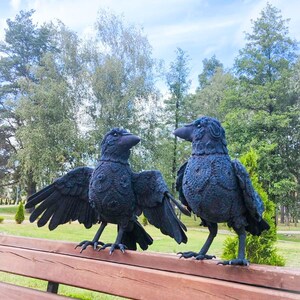 May include: Two black raven sculptures with detailed feather textures stand on a wooden bench. One raven has its wings spread, while the other stands upright. The background features trees and a blue sky with clouds.