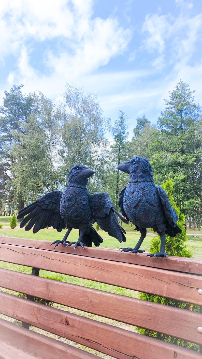 May include: Two black raven sculptures with detailed feather textures stand on a wooden bench. One raven has its wings spread, while the other stands upright. The background features trees and a blue sky with clouds.