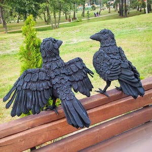 May include: Two black raven sculptures with detailed feather textures are perched on a wooden bench. One raven has its wings spread wide, while the other stands upright. The background features a park with green grass and trees.
