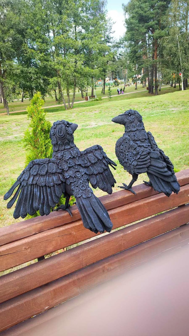 May include: Two black raven sculptures with detailed feather textures are perched on a wooden bench. One raven has its wings spread wide, while the other stands upright. The background features a park with green grass and trees.