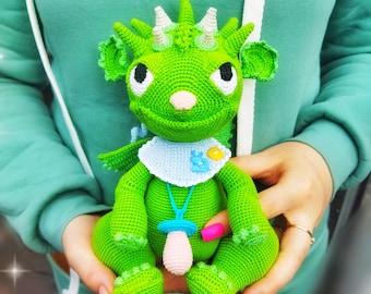 May include: A green crocheted baby dragon amigurumi toy wearing a blue bib with a pacifier. The toy has a white horn and black eyes. The caption reads "BABY DRAGON AMIGURUMI PATTERN".