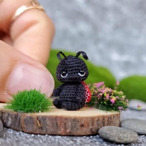 May include: A small black crocheted ladybug with red and white spots on its back. The ladybug has a sad expression and is sitting on a wooden surface with green moss and small pink flowers.