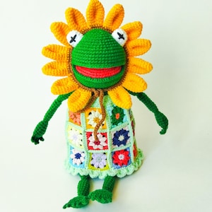 May include: A green crocheted frog wearing a yellow crocheted sunflower hat and a multi-colored crocheted dress with a flower pattern.
