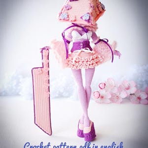 May include: A crocheted doll featuring a large pyramid-shaped head adorned with small flowers, wearing a white top, purple corset, and ruffled skirt. The doll holds a large rectangular blade and wears purple shoes. Text reads: Crochet pattern pdf in english.