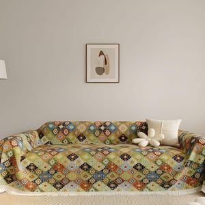 May include: A sofa draped with a patterned throw blanket. The blanket displays a geometric design in green, orange, blue, and yellow. A white cushion and a flower-shaped cushion are on the sofa. The sofa is in a living room.