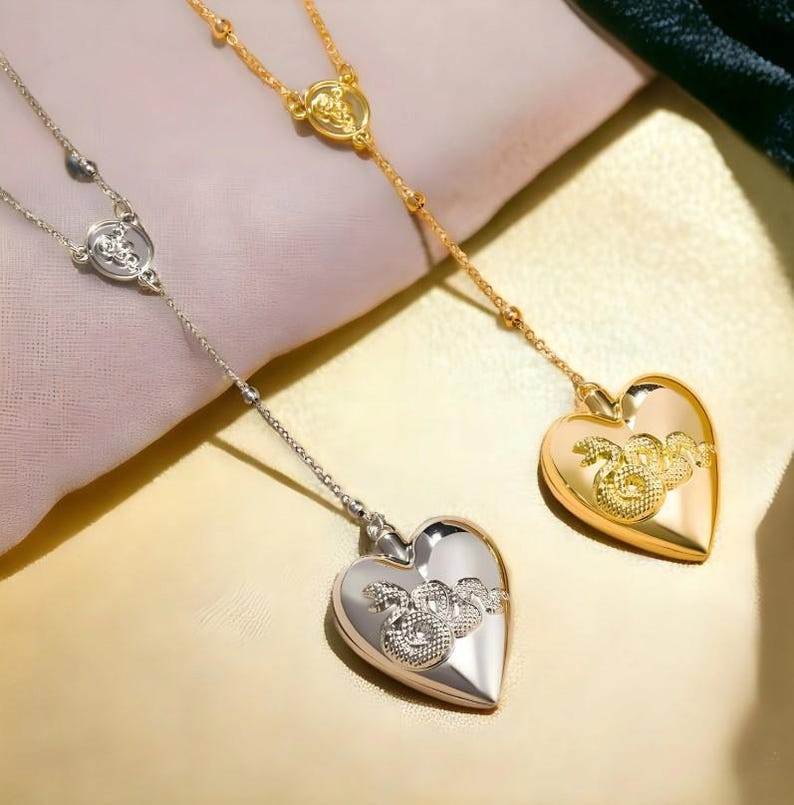 May include: Two silver and gold heart-shaped locket necklaces with a snake design on the front. The necklaces are on a white background.