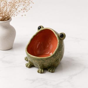 Peut inclure: Un bol en céramique en forme de grenouille avec un extérieur vert olive moucheté et un intérieur orange vif et lisse. La grenouille présente deux yeux noirs saillants sur le dessus et quatre petites pattes à la base, formant un récipient ouvert.