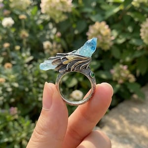 May include: A silver ring featuring a textured band with a small green stone accent and a large, light blue, raw crystal point set within a decorative, wing-like metal structure that extends horizontally across the top of the ring band.