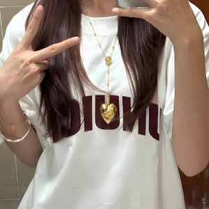 May include: A white t-shirt with the word "DIOR" printed in maroon on the front. A gold chain necklace with a heart-shaped pendant hangs from the neck.