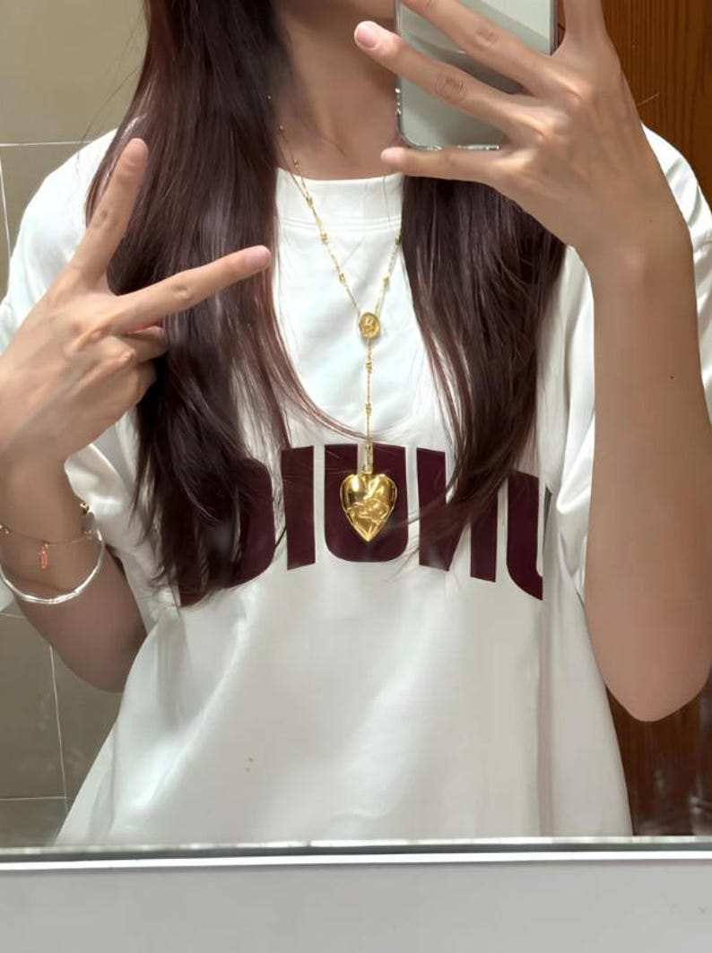 May include: A white t-shirt with the word "DIOR" printed in maroon on the front. A gold chain necklace with a heart-shaped pendant hangs from the neck.