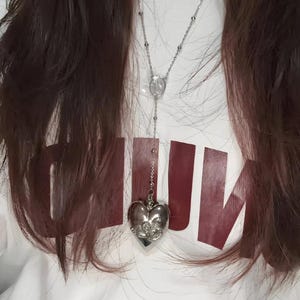 May include: A silver heart-shaped pendant necklace with a delicate chain. The pendant has a detailed design and is hanging from a white shirt with the word "RUN" printed in maroon.