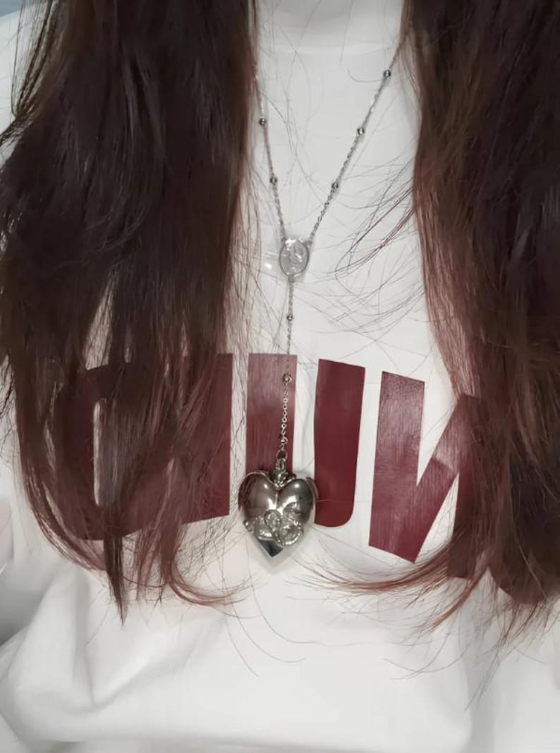 May include: A silver heart-shaped pendant necklace with a delicate chain. The pendant has a detailed design and is hanging from a white shirt with the word "RUN" printed in maroon.