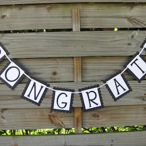 K&ouml;nnte beinhalten: Ein Banner mit dem Wort "CONGRATS" in schwarzen Buchstaben auf wei&szlig;en Quadraten mit einem geschwungenen schwarzen Rand. Das Banner ist an einem wei&szlig;en Band aufgereiht und h&auml;ngt vor einem Holzzaun.