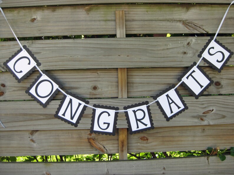K&ouml;nnte beinhalten: Ein Banner mit dem Wort "CONGRATS" in schwarzen Buchstaben auf wei&szlig;en Quadraten mit einem geschwungenen schwarzen Rand. Das Banner ist an einem wei&szlig;en Band aufgereiht und h&auml;ngt vor einem Holzzaun.