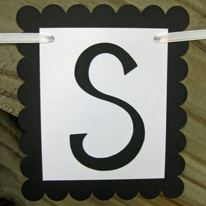 K&ouml;nnte beinhalten: Ein Banner mit einem wei&szlig;en rechteckigen Mittelteil mit dem schwarzen Buchstaben "S". Das Banner ist von einem schwarzen, gewellten Rand umrahmt und hat ein wei&szlig;es Band zum Aufh&auml;ngen. Der Hintergrund ist eine Holzoberfl&auml;che.