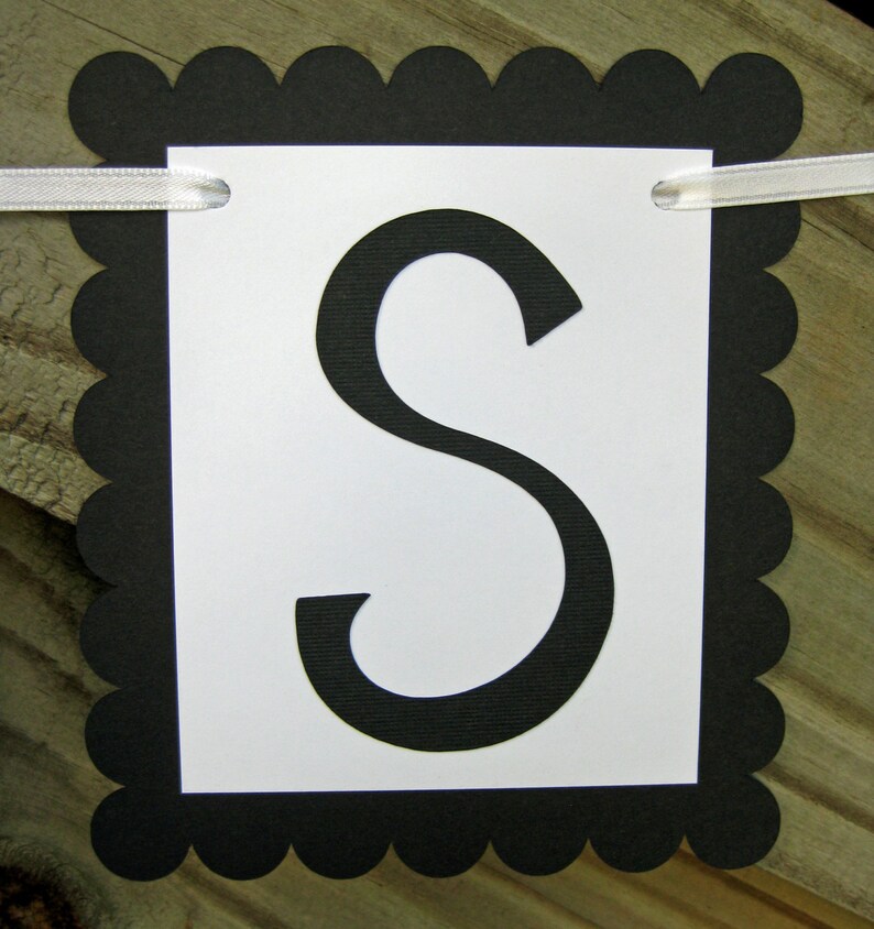 K&ouml;nnte beinhalten: Ein Banner mit einem wei&szlig;en rechteckigen Mittelteil mit dem schwarzen Buchstaben "S". Das Banner ist von einem schwarzen, gewellten Rand umrahmt und hat ein wei&szlig;es Band zum Aufh&auml;ngen. Der Hintergrund ist eine Holzoberfl&auml;che.