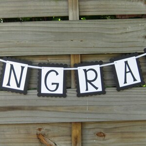 K&ouml;nnte beinhalten: Ein Banner mit dem Wort "CONGRATS" in schwarzen Buchstaben auf wei&szlig;en rechteckigen Flaggen mit einem geschwungenen schwarzen Rand. Das Banner ist an einem wei&szlig;en Band aufgereiht und h&auml;ngt vor einem Holzzaun.