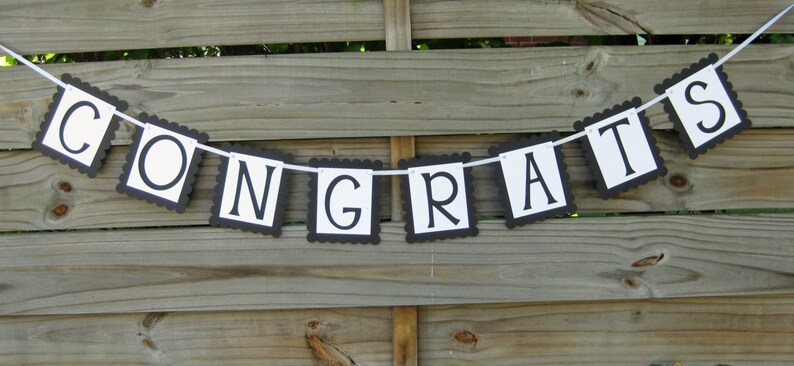 K&ouml;nnte beinhalten: Ein Banner mit dem Wort "CONGRATS" in schwarzen Buchstaben auf wei&szlig;en rechteckigen Flaggen mit einem geschwungenen schwarzen Rand. Das Banner ist an einem wei&szlig;en Band aufgereiht und h&auml;ngt vor einem Holzzaun.