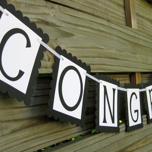 K&ouml;nnte beinhalten: Ein Banner mit dem Wort "CONGRATS" in schwarzen Gro&szlig;buchstaben auf wei&szlig;en quadratischen Flaggen mit einem schwarzen, gewellten Rand. Das Banner ist an einem wei&szlig;en Band aufgereiht und h&auml;ngt vor einem Holzzaun.