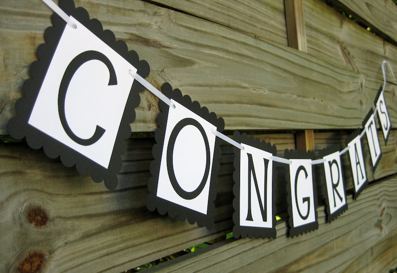 K&ouml;nnte beinhalten: Ein Banner mit dem Wort "CONGRATS" in schwarzen Gro&szlig;buchstaben auf wei&szlig;en quadratischen Flaggen mit einem schwarzen, gewellten Rand. Das Banner ist an einem wei&szlig;en Band aufgereiht und h&auml;ngt vor einem Holzzaun.