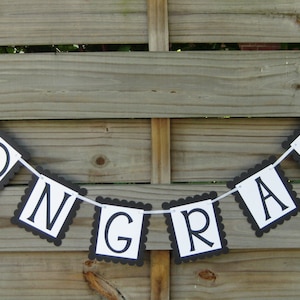 K&ouml;nnte beinhalten: Ein Banner mit der Aufschrift "CONGRATS" in schwarzen Buchstaben auf wei&szlig;en Quadraten mit einem schwarzen, gewellten Rand. Das Banner ist an einer wei&szlig;en Schnur befestigt und h&auml;ngt vor einem Holzzaun.