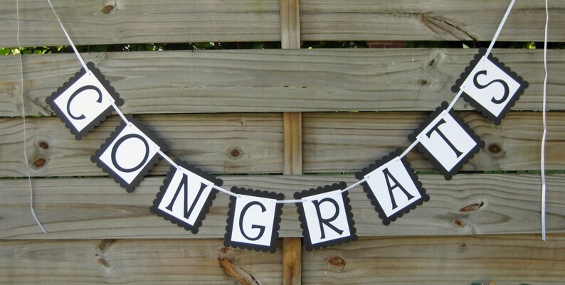 K&ouml;nnte beinhalten: Ein Banner mit der Aufschrift "CONGRATS" in schwarzen Buchstaben auf wei&szlig;en Quadraten mit einem schwarzen, gewellten Rand. Das Banner ist an einer wei&szlig;en Schnur befestigt und h&auml;ngt vor einem Holzzaun.