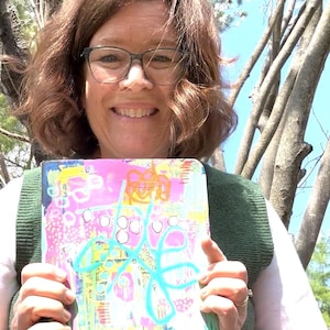 May include: A colorful journal with abstract art in pink, yellow, blue, and orange. The cover features a turquoise floral design and various shapes. The journal is held in front of a person wearing glasses and a green vest.