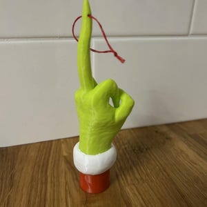 May include: A lime green hand ornament with a middle finger extended, featuring a white cuff and a red sleeve base. A thin red string is attached to the top of the middle finger for hanging. The hand has a textured surface and a distinct thumb.