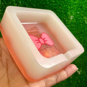 Puede incluir: Cenicero cuadrado con un diseño de lazo rosa en el interior. El cenicero está hecho de un material translúcido con un borde color melocotón. El lazo es de color rosa brillante. El cenicero se sostiene en una mano.