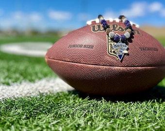 Może przedstawiać: Brązowo-biała piłka nożna z nadrukiem "Wilson" i "Junior Size". Na piłce leży naszyjnik z koralików z zawieszką Baltimore Ravens.