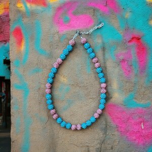 May include: A necklace made of alternating blue and pink round beads. The necklace is made of small, sparkly beads and is hanging against a colorful, textured wall.