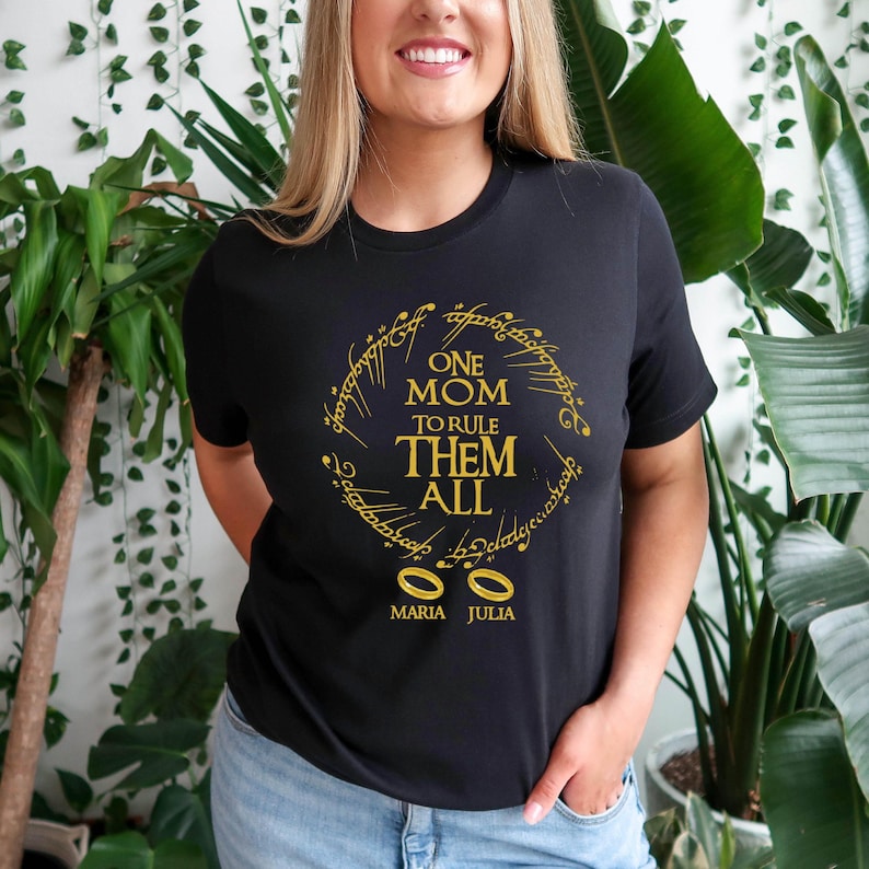 May include: Black t-shirt with gold text that reads "ONE MOM TO RULE THEM ALL" in a circular design, with two gold rings below. The name "MARIA JULIA" is printed below the rings.
