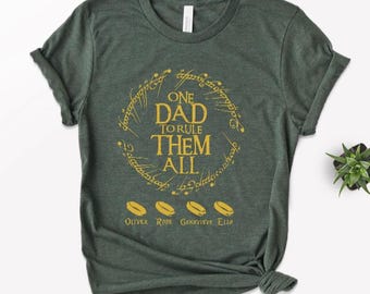 May include: Dark green t-shirt with a gold design. The design includes the text "ONE DAD TO RULE THEM ALL" in a circular pattern, with four gold rings below and names: Oliver, Rose, Genevieve, and Ella.