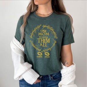 May include: Green t-shirt with the text "ONE MOM TO RULE THEM ALL" in gold lettering, surrounded by a ring design. Below the text are two gold rings and the names "MARIA JULIA".