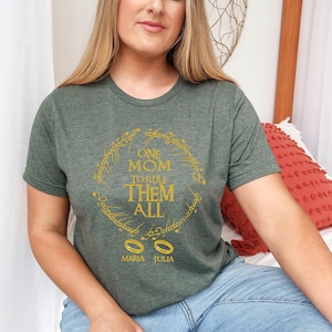 May include: A green t-shirt with a gold design featuring the text "ONE MOM TO RULE THEM ALL" in a circular pattern. Below the text are two gold rings and the names "MARIA JULIA". The shirt is short-sleeved.
