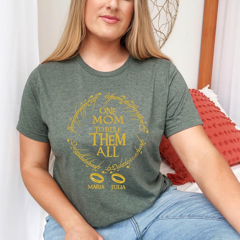 May include: A green t-shirt with a gold design featuring the text "ONE MOM TO RULE THEM ALL" in a circular pattern. Below the text are two gold rings and the names "MARIA JULIA". The shirt is short-sleeved.