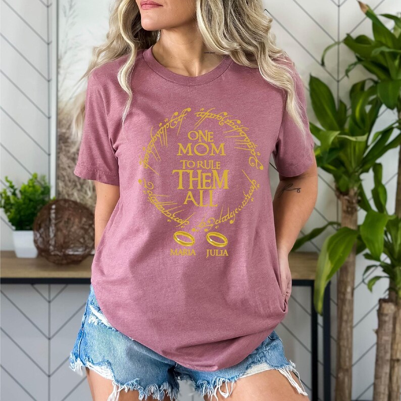 May include: A mauve t-shirt with the text "ONE MOM TO RULE THEM ALL" in a gold ring design. Below the ring are two gold rings and the names "MAMA JULIA". The shirt has short sleeves.