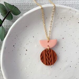 May include: A gold chain necklace with a brown and pink clay pendant. The pendant is made up of a circle with a textured design and a half-circle.
