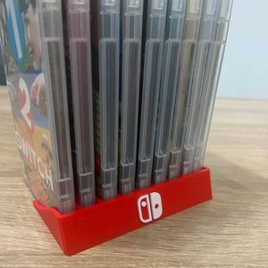 Peut inclure: Un support de cartouches de jeu en plastique rouge avec le logo Nintendo Switch en blanc. Le support est rempli de plusieurs boîtiers de jeu transparents. Le support repose sur une surface en bois brun clair.