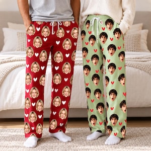May include: Two pairs of lounge pants with drawstring waistbands. The left pair is red with a repeating pattern of white hearts and a person's face. The right pair is light green with a repeating pattern of red hearts and a person's face.