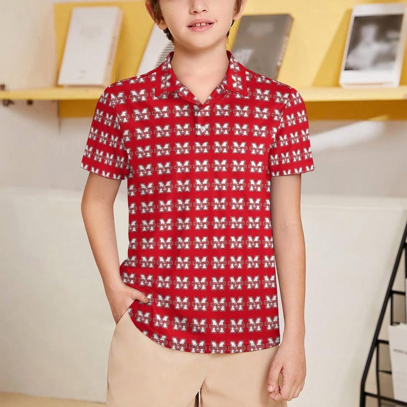 May include: A red short-sleeved button-down shirt with a repeating white and red "M" pattern. The shirt is worn by a person and paired with khaki-colored pants. The shirt has a classic collar and a relaxed fit.