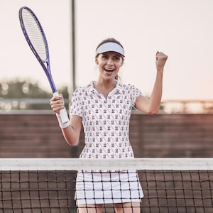 K&ouml;nnte beinhalten: Eine Frau tr&auml;gt ein wei&szlig;es Tennisshirt mit einem rot-wei&szlig;-blauen Muster. Sie h&auml;lt einen Tennisschl&auml;ger und l&auml;chelt. Sie steht auf einem Tennisplatz.