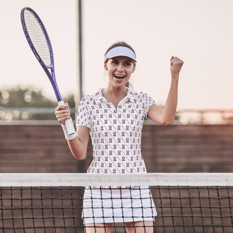 K&ouml;nnte beinhalten: Eine Frau tr&auml;gt ein wei&szlig;es Tennisshirt mit einem rot-wei&szlig;-blauen Muster. Sie h&auml;lt einen Tennisschl&auml;ger und l&auml;chelt. Sie steht auf einem Tennisplatz.