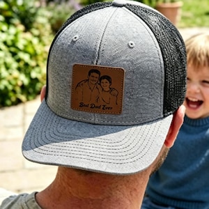 Può includere: Cappellino da camionista grigio e nero con una toppa in pelle marrone. La toppa presenta un disegno di un uomo e un bambino con la scritta "Best Dad Ever". Il cappellino ha una visiera curva e un retro in rete.